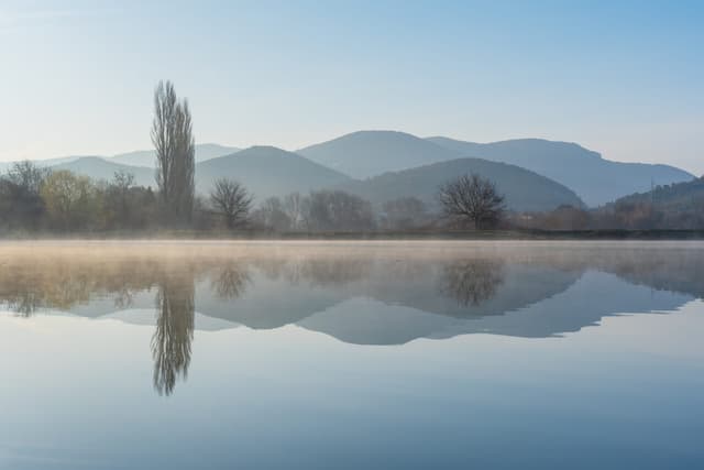 A still lake mirroring hills and bare trees in mist